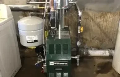 Green Williamson furnace in a basement, connected to pipes and a water heater, with a washing machine nearby.