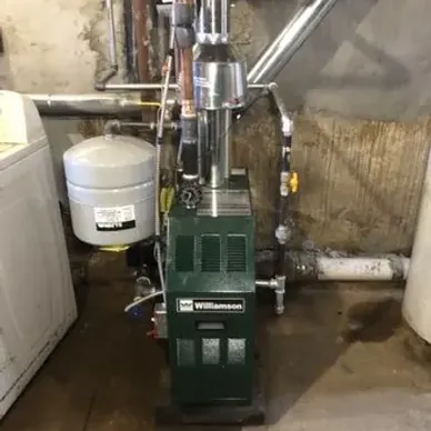 Green Williamson furnace in a basement, connected to pipes and a water heater, with a washing machine nearby.