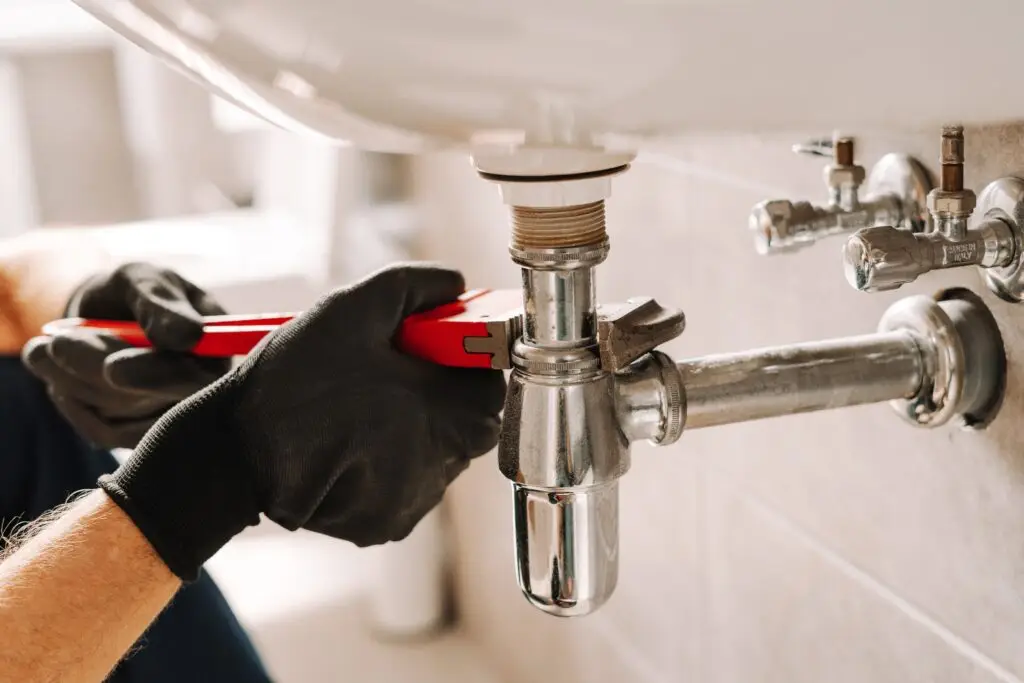 Plumber using a red wrench to tighten a pipe under a sink, wearing black gloves.