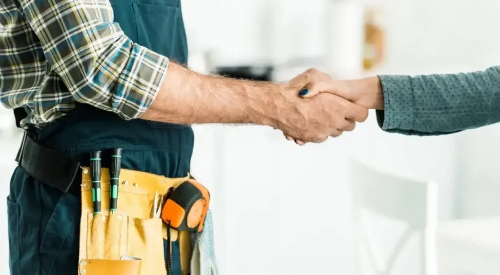 Plumber in plaid shirt and tool belt shaking hands with a person in a blue sweater.